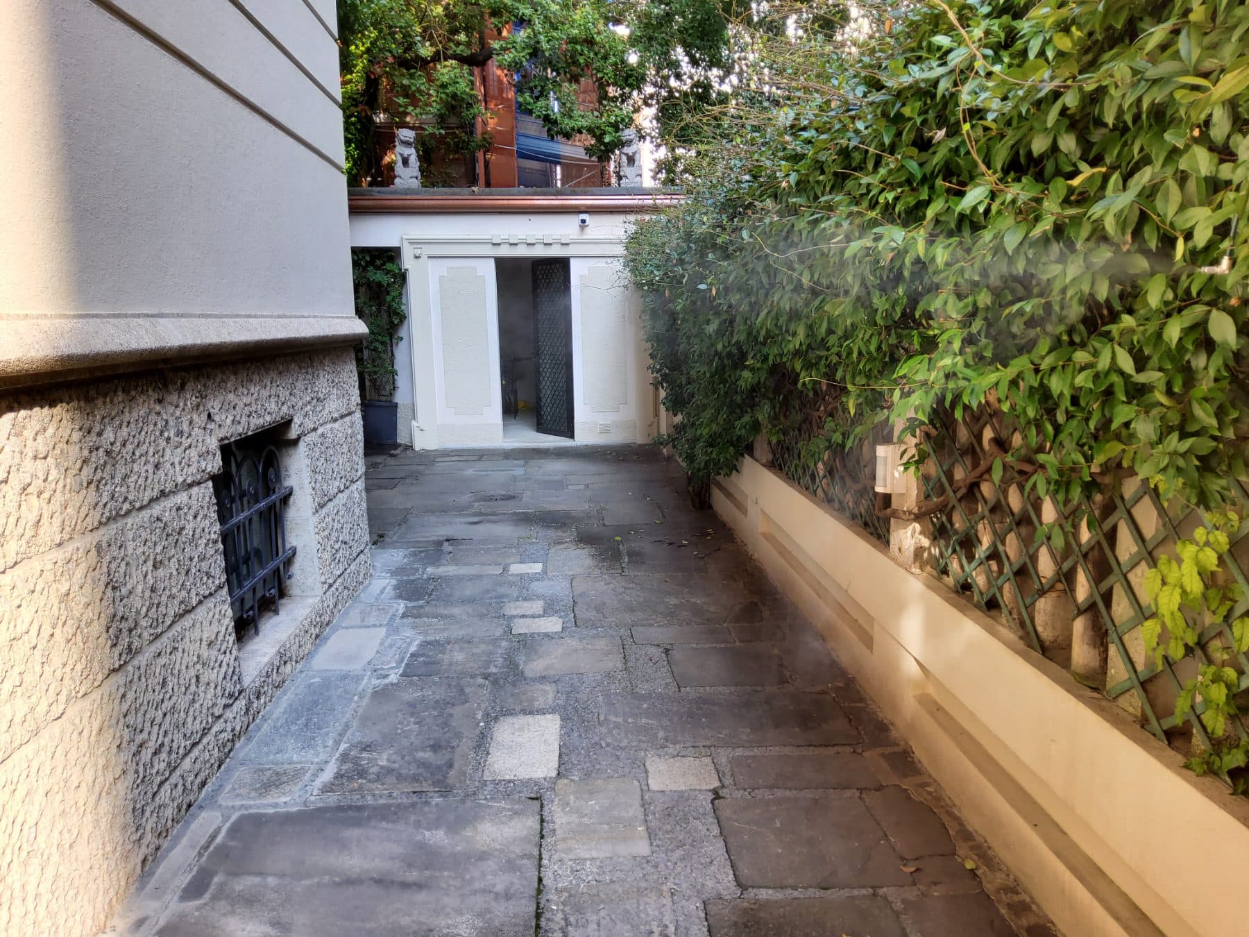 Passaggio pavimentato in pietra tra il palazzo e una siepe, con la nebulizzazione lungo il muro